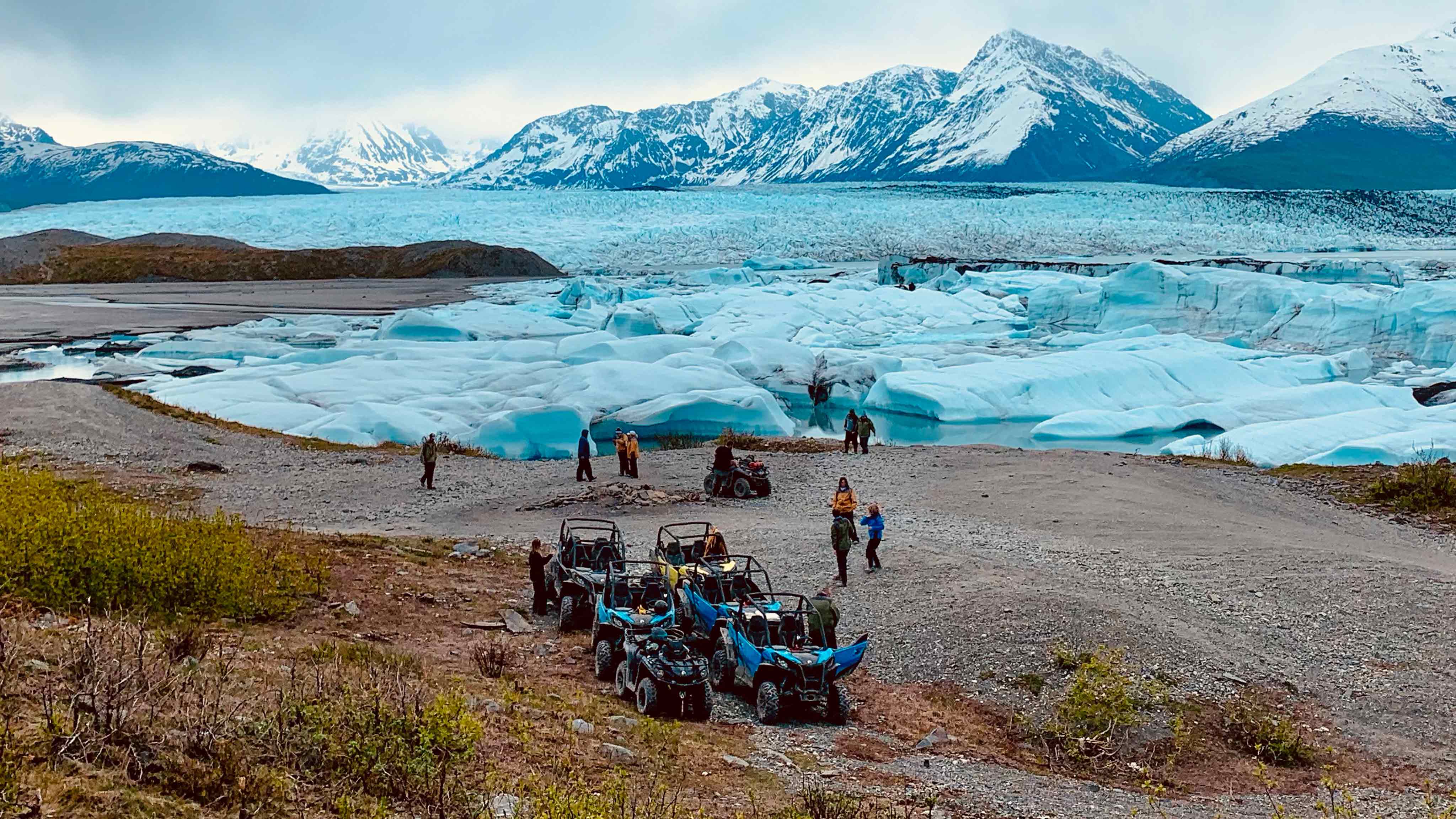 People gather near water and mountains with off-road vehicles