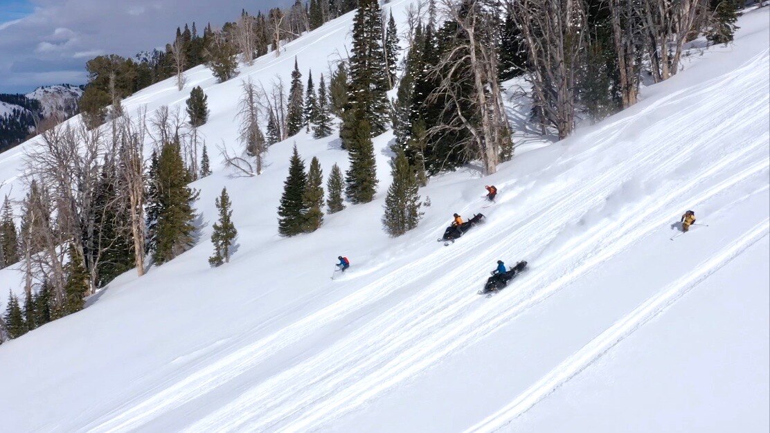 People going down hill on snowmobiles and skis