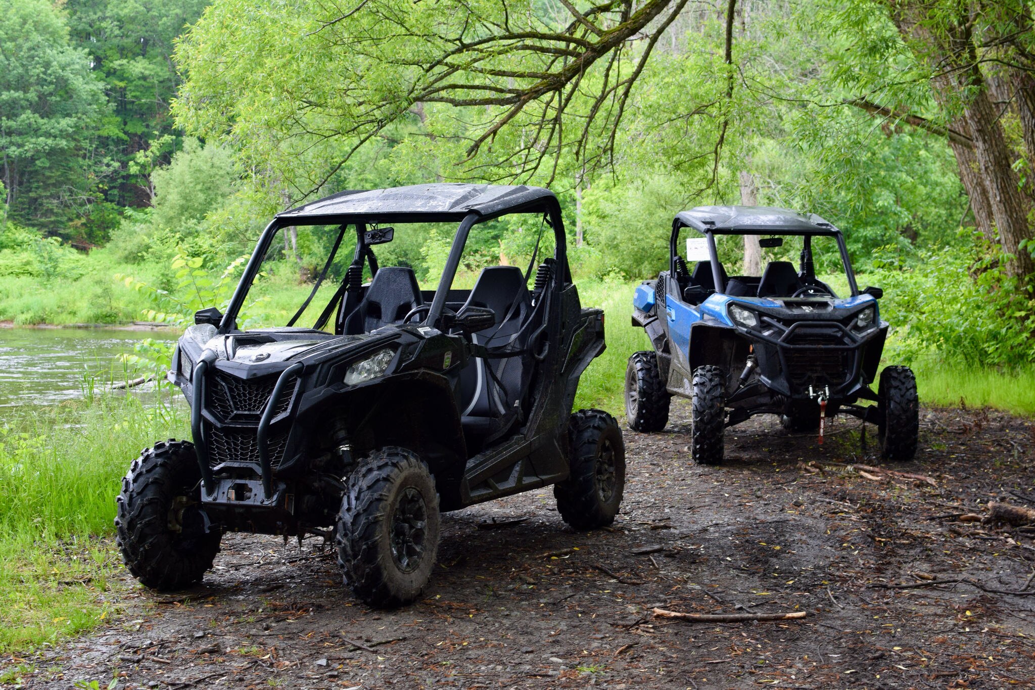 Two Can-Am Side-by-Sides parked on dirt, next to flowing water, under trees