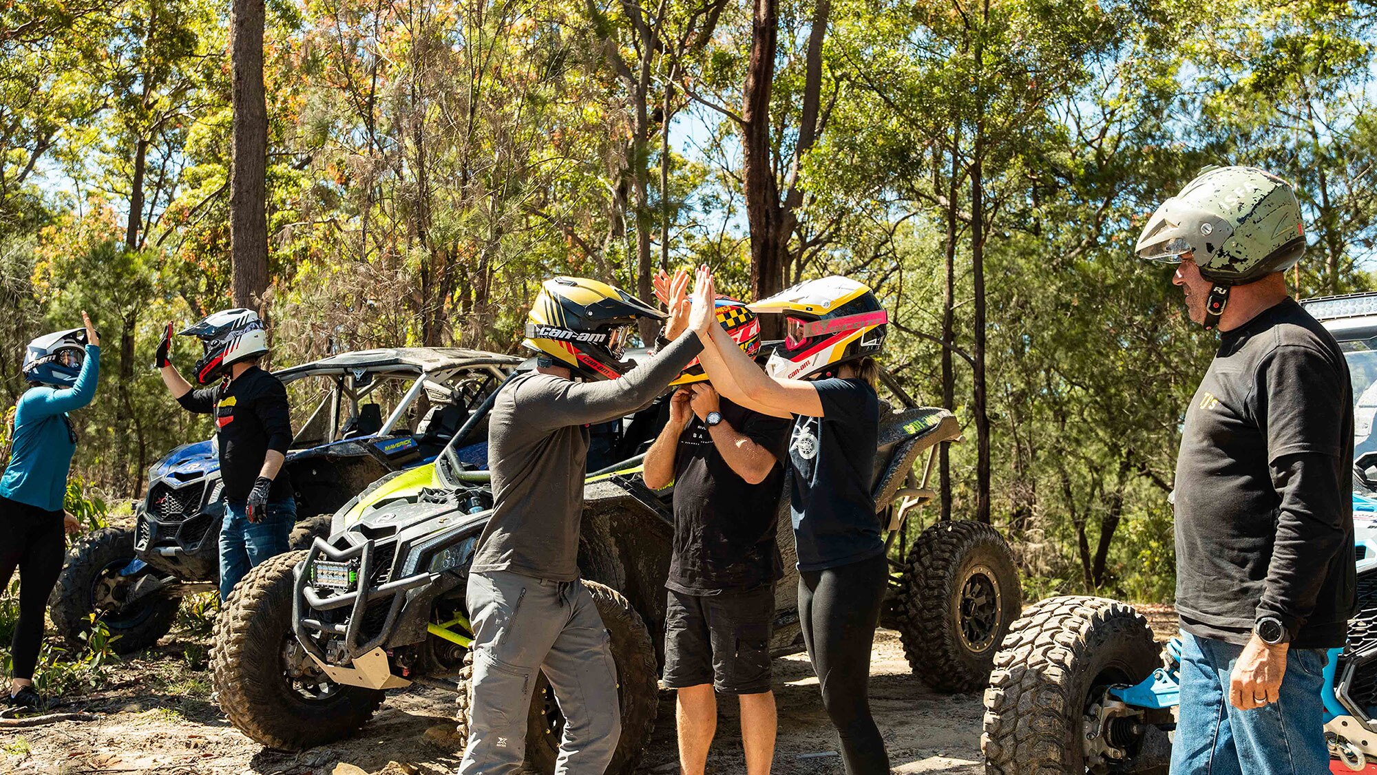 A group of people high fiving and celebrating in front of Can-Am side by sides with trees in the back.