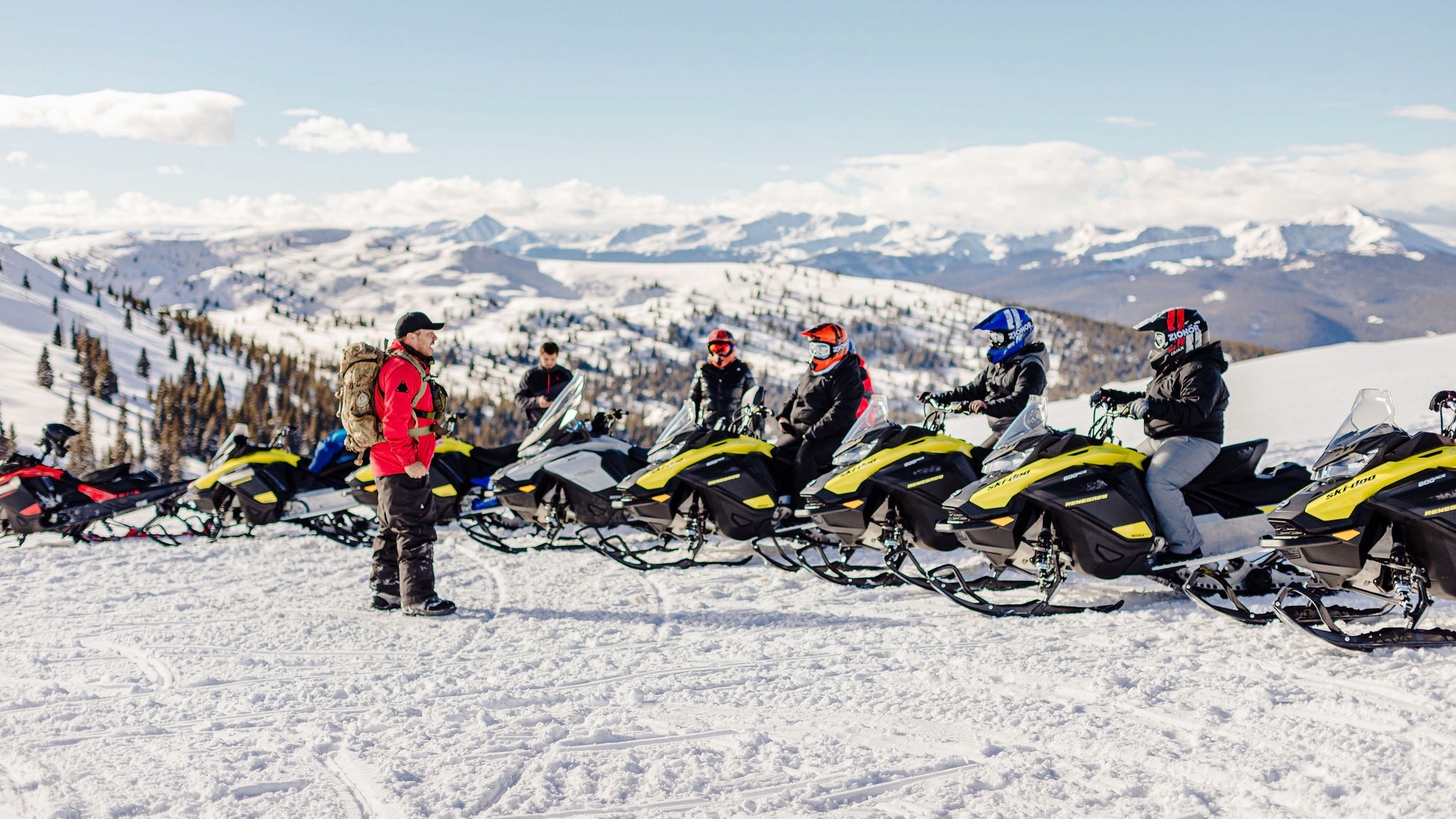 A group of snowmobile riders on a mountain top listening to the guide.