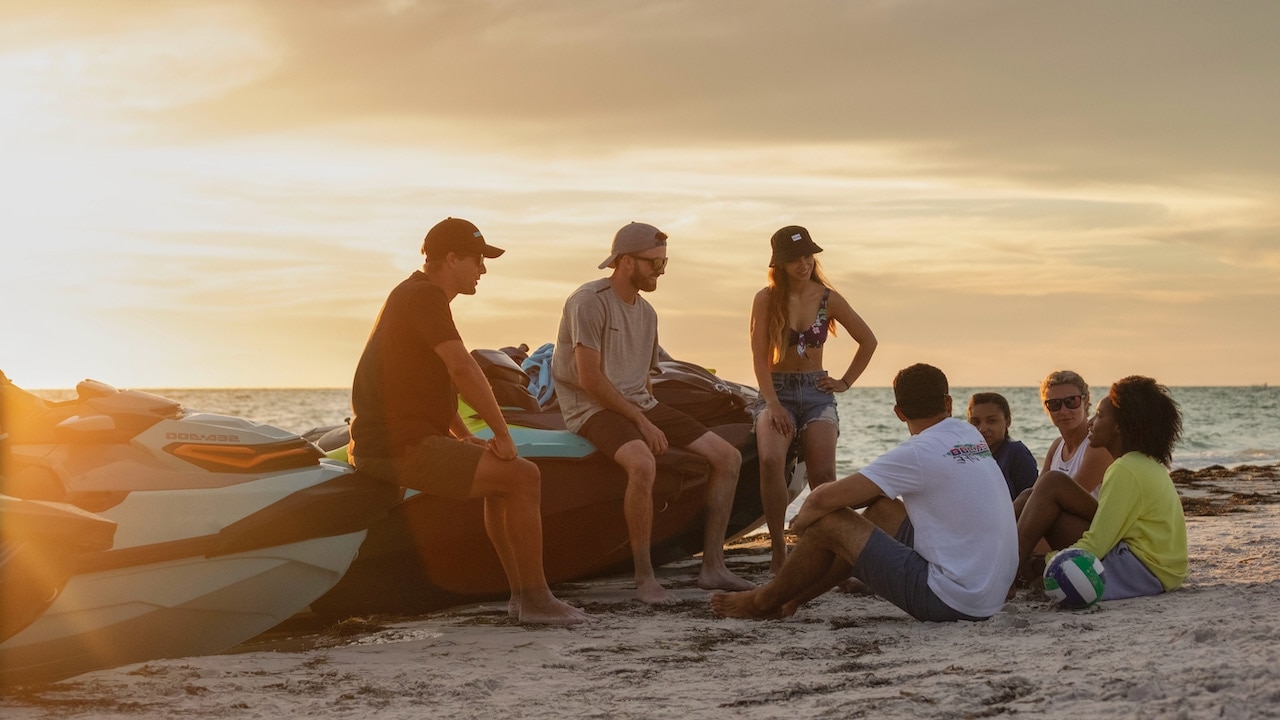 A group of people sitting by the water on the sand, leaning on docked Sea-doos, laughing and smiling