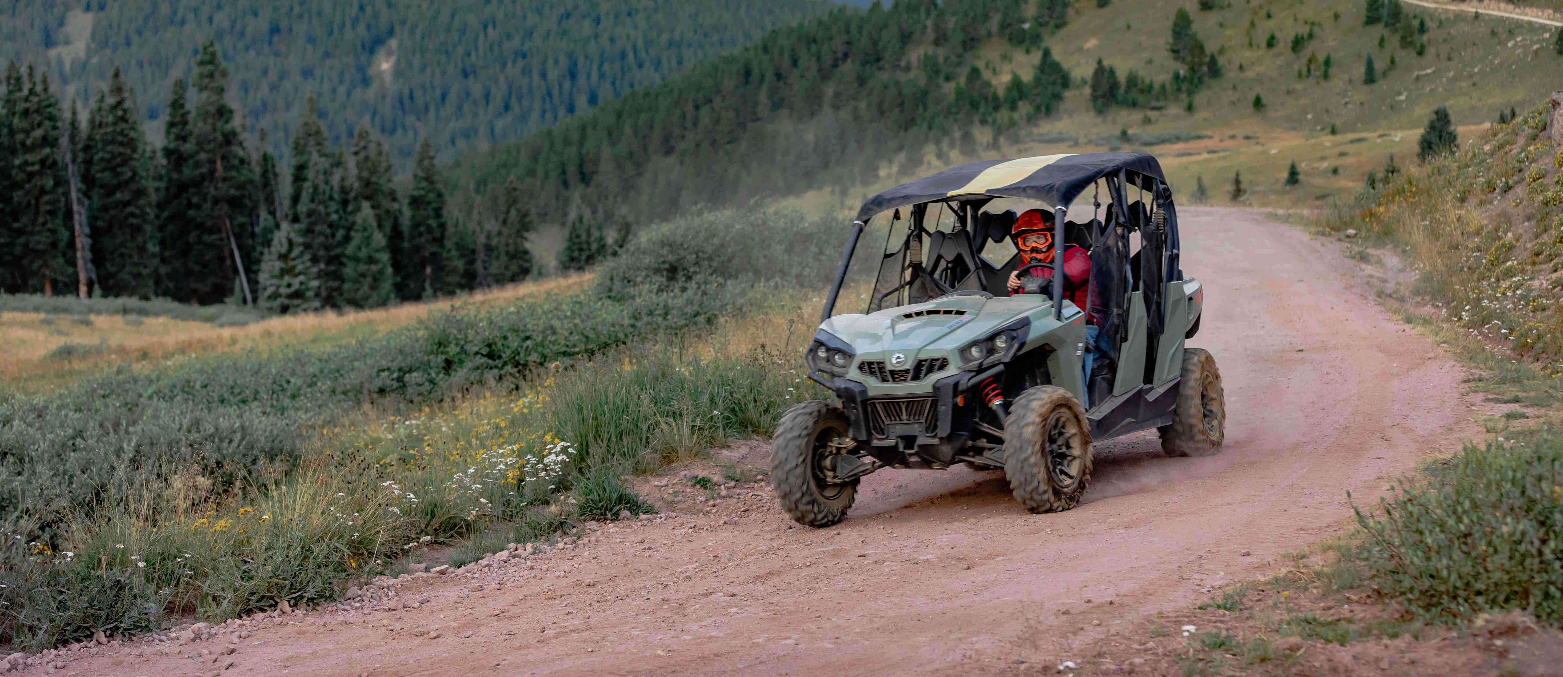 A person riding a Can-Am side-by-side on a trail through the mountains with trees and a blue sky in the back 