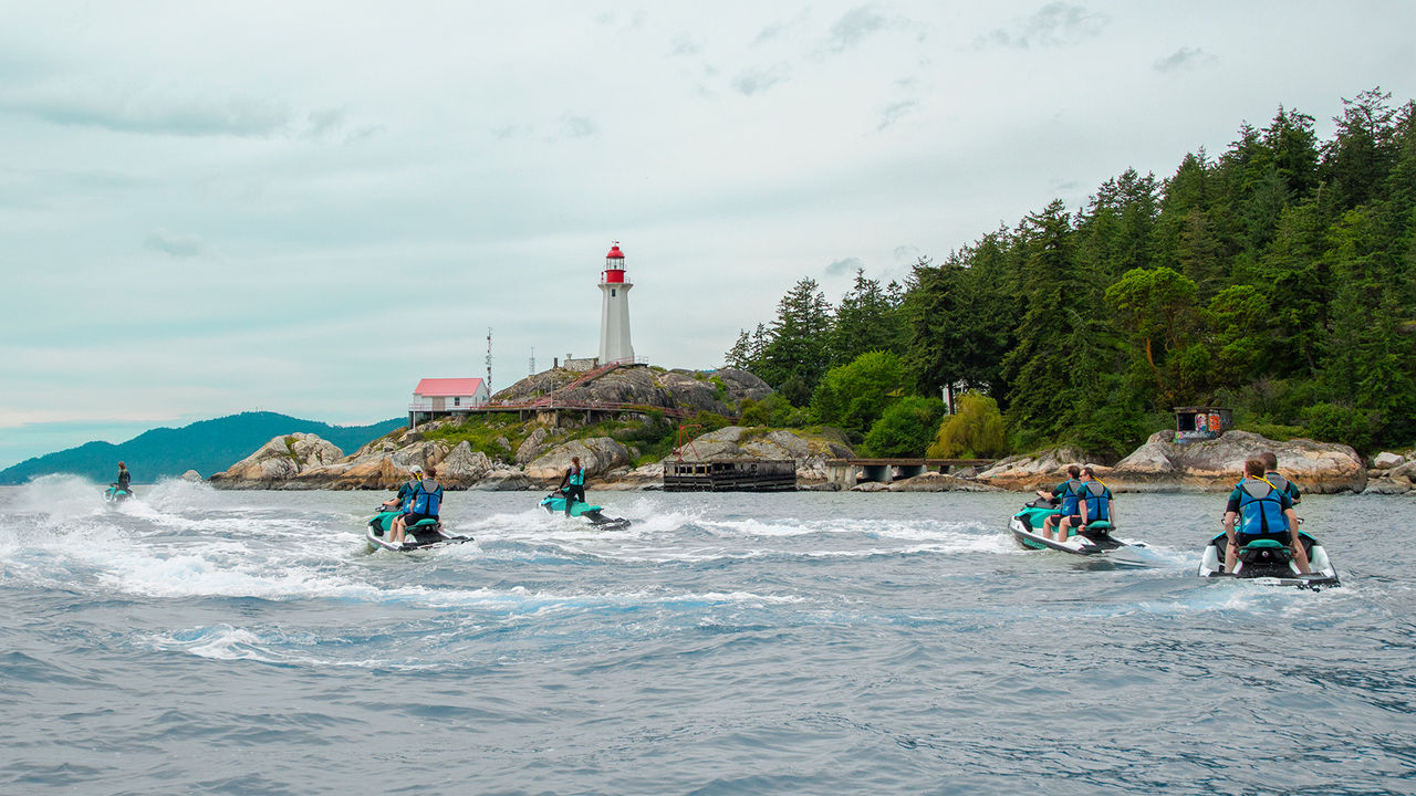 A group of people on Sea-Doos, with a lighthouse and mountains in the back